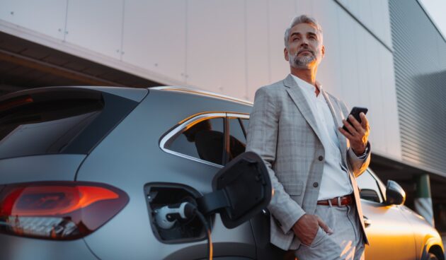 Businessman holding smartphone while charging car at electric vehicle charging station, closeup.